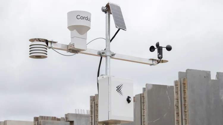 Wetterstation von Cordulus auf einem Mast an einer Baustelle, dahinter gestapelte Betonfertigteile und Schalungselemente